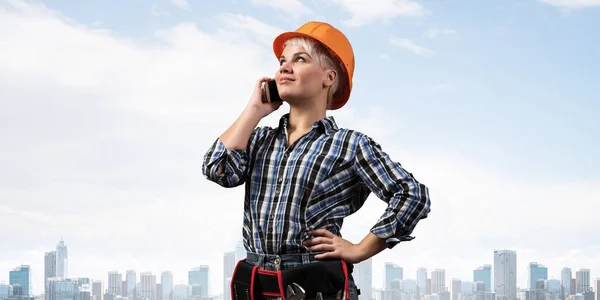 迷人的金发女人穿着工作服 头戴安全帽 在智能手机上聊天 穿着格子蓝色衬衫站在城市景观背景上的年轻工人的画像 商务沟通和对话 — 图库照片