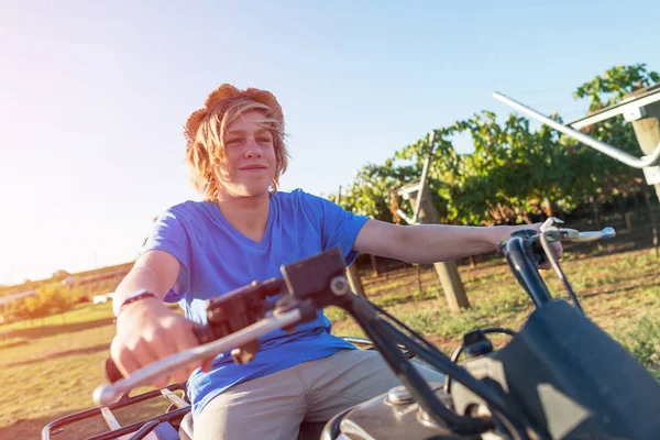 Young farmer riding quad bike on background of vine rows. Traditional farming in vineyard. Happy blond guy in straw hat and and blue t-shirt sitting on bike. Ecological food production in countryside.