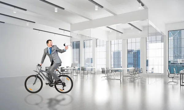 Man in business suit riding bicycle at conference hall. Businessman with megaphone looking back on bike at loft office interior with panoramic windows. Business presentation and announcement.