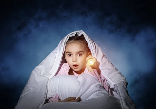被手电筒藏在毯子下的惊恐女孩 惊慌失措的孩子躺在家里的床上 儿童夜惊小女孩晚上睡不着 深蓝色天空背景穿着睡衣的孩子 — 图库照片