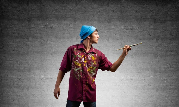 Young artist gesturing with paintbrush. Male painter in dirty shirt and bandana standing on grey wall background. Creative hobby and artistic occupation. Art classes concept with copy space.