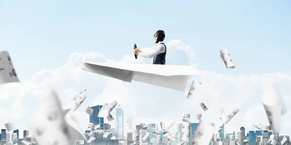 飛行士の帽子とゴーグルのビジネスマンは 都市景観の上に紙飛行機を運転します 雲と青空を背景に落ちる紙 大きな紙飛行機で楽しむ表情パイロット — ストック写真