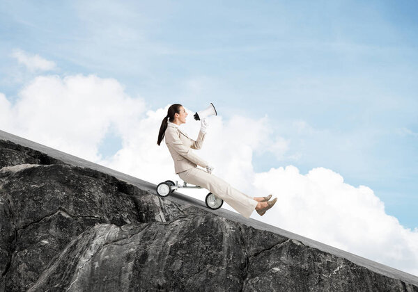 Woman shouting into megaphone and riding downhill on road. Young employee in white business suit going down on small bike outdoor. Fast business startup. Advertising and promotion concep