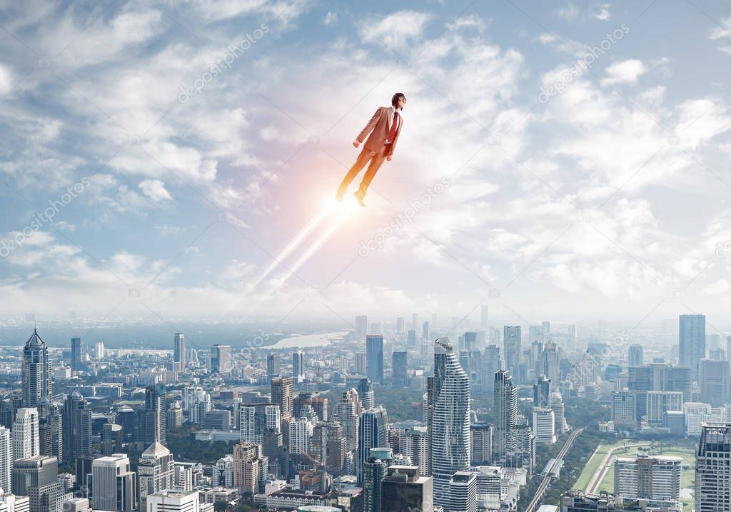 Businessman in suit and aviator hat flying in blue sky as superhero. Corporate manager as superman launching upwards with jet flame above modern city. Successful business startup motivation concept