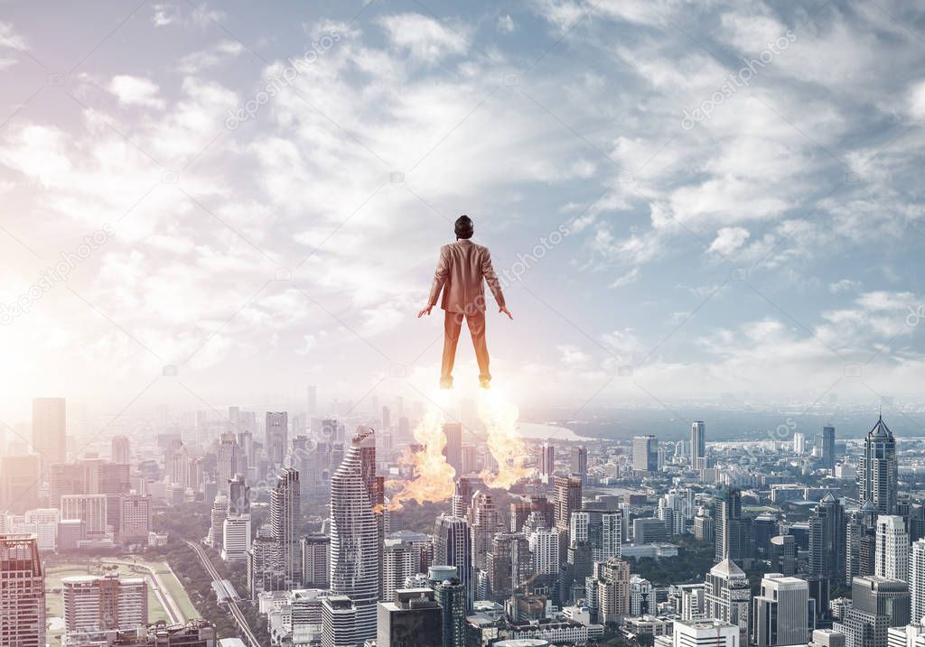 Businessman in suit and aviator hat flying in blue sky as superhero. Back view of corporate manager as superman launching upwards with jet flame above modern city. Successful business startup