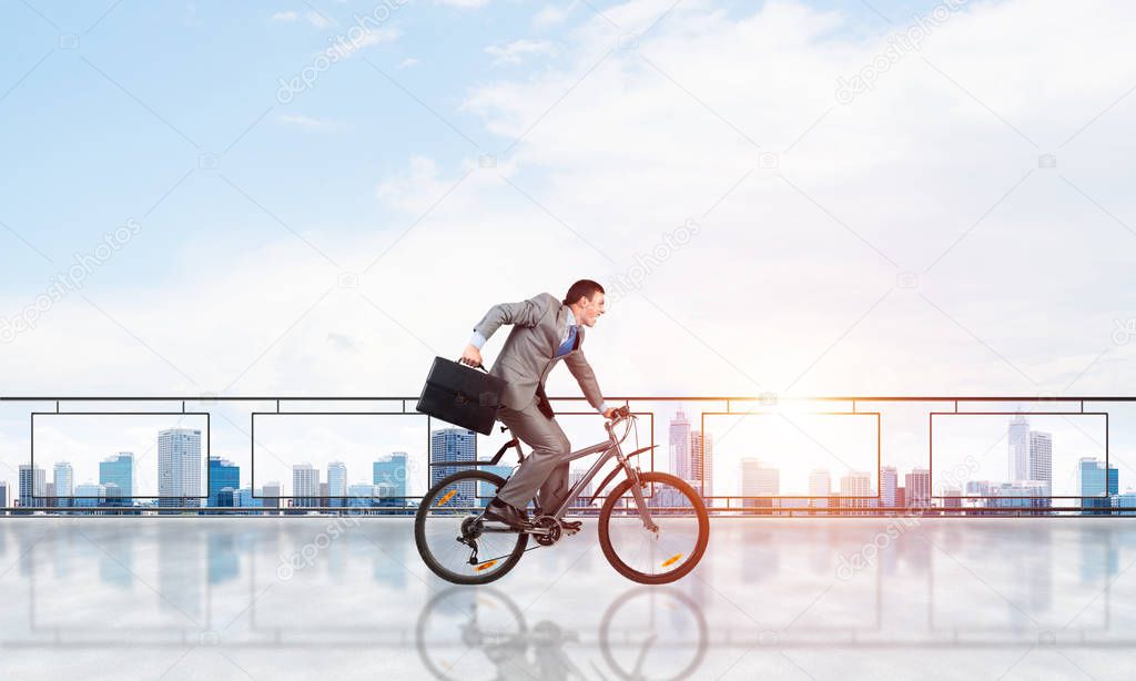 Man wearing business suit riding bicycle on penthouse balcony. Young cyclist with suitcase on background of business district. Terrace with modern downtown view. Time management and business activity