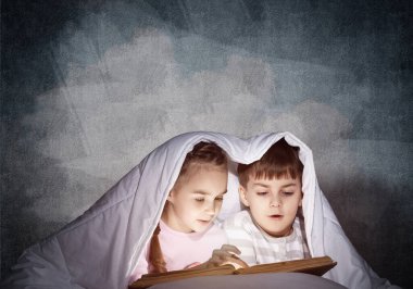 Dalmış küçük kız ve erkek yatmadan sonra yatakta kitap okuyor. Güzel çocuklar battaniyenin altında saklanıyorlar. Pijamalı okul öncesi çocuklar peri masalları okuyor. Gri duvar arka planlı yatak odası.