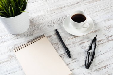 Bir fincan kahveyle ofis çalışma alanı. Düz tahta masa, açık spiral defter, gözlük ve kalemle. Kahve molası. Notlar için boş defter sayfası. Çaydanlıkta yeşil bitki.