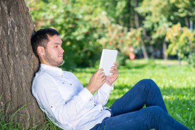 Güneşli bir günde parkta ağacın altında tablet bilgisayar kullanan genç bir adam. Tablet ekranına bakan yakışıklı bir iş adamı. Ağaca yaslanmış yeşil çimlerde oturan sakin bir adam. Mobil iletişim konsepti.