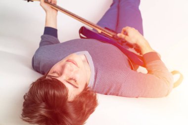 Young man with closed eyes practicing electric or bass guitar. Guitarist lying back on white sofa. Close-up caucasian musician in sweater and jeans playing acoustic guitar. Playing musical instruments clipart