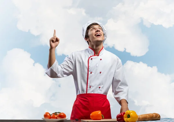 年轻的男厨师站在那里 手指向上指向 漂亮的厨师戴着白帽 蓝天背景的红围裙 餐厅广告 食品零售活动公告 饮食服务 — 图库照片
