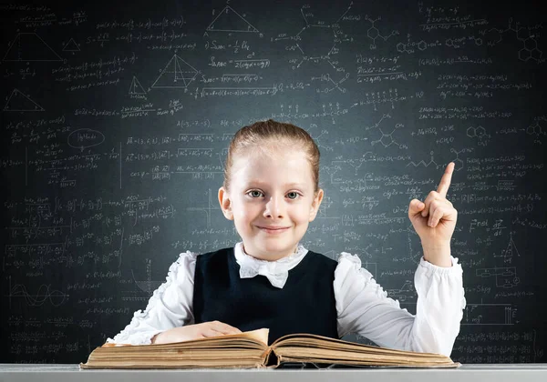 机に座って本を開いて微笑む少女 学校での研究と教育 学校の幸せな女子高生は指差して指を振る 賢い女の子読書本上の黒板の背景 — ストック写真