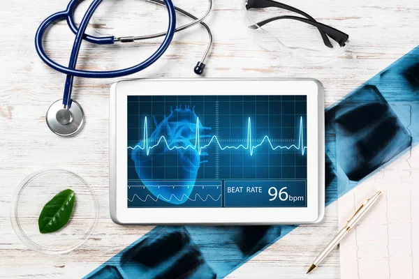 Medical diagnostics in modern hospital. Tablet computer with medical application interface on screen. Top view x-ray images, stethoscope and cardiogram on wooden desk. Digital technology in clinic.