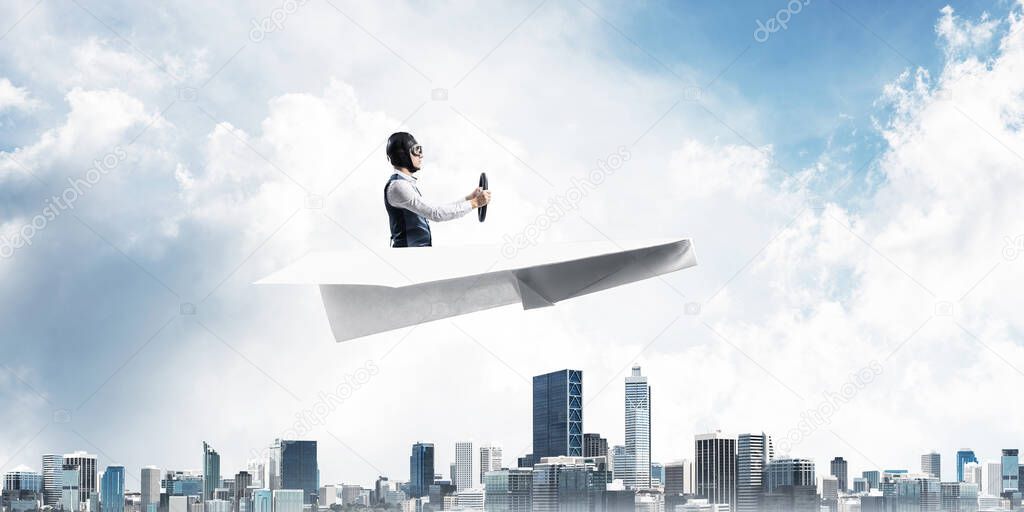 Business motivation concept with pilot sitting in paper airplane. Funny man in aviator hat and goggles driving paper plane above city. Dreaming to fly in sky. Modern megapolis with high buildings.