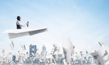 Havacı bulutlu mavi gökyüzünde iş merkezi üzerinde kağıt uçak sürüş. Gökdelenlerin arka planına düşen kağıtlar. Deri kasklı pilot kağıt düzlemde otururken ve direksiyonu tutarak.