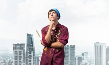 Düşünceli ressam modern şehir manzarası arka planında kollarını kavuşturmuş duruyor. Hayallere dalmış genç bir adamın portresi. Sanatsal meslek ve yaratıcı meslek. Sanat okulu öğrencisi pozu.