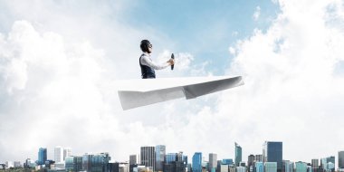 Kağıt uçakta oturan pilot ile iş liderliği ve motivasyon konsepti. Havacı şapkalı komik adam ve şehrin üzerinde kağıt uçak süren gözlükler. Yüksek binalarile modern megapolis.