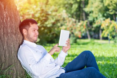 Güneşli bir günde parktaki ağacın altında tablet bilgisayar kullanan bir adam. Tablet ekranına bakan yakışıklı adam. Ağaçlara yaslanmış yeşil çimlerde oturan mutlu adam. Dijital teknoloji ve iletişim