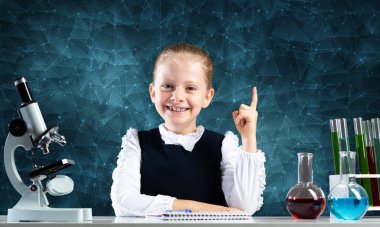 İlköğretim fen sınıfında masa başında oturan küçük bir bilim kadını. Okulda araştırma ve eğitim. Mikroskop, matara ve test tüplü kimya laboratuvarı. Mutlu liseli kız işaret ediyor