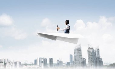 Havacı şapkalı işadamı ve gözlüklü, şehir merkezinin üzerinde yüksek binalarla kağıt uçak sürüyor. Mavi gökyüzü ile modern megalopolis panorama. Karışık medya iş kavramı. Hayal ve hayal gücü.