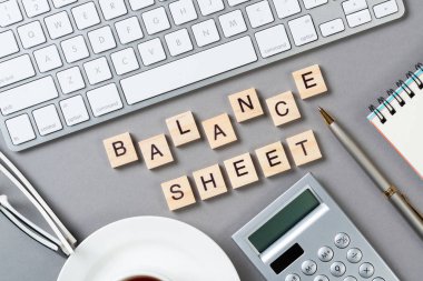 Tahta küplerdeki harflerle denge tablosu kavramı. Malzemelerle dolu ofis hayatı. Düz gri yüzey, bilgisayar klavyesi ve hesap makinesiyle yatıyordu. Bütçe planlaması, denetim ve muhasebe.