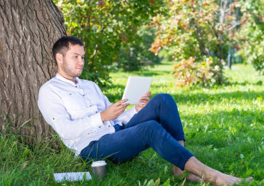 Güneşli bir günde parkta ağacın altında tablet bilgisayar kullanan genç bir adam. Tablet ekranına bakan yakışıklı bir iş adamı. Ağaca yaslanmış yeşil çimlerde oturan sakin bir adam. Mobil iletişim konsepti.