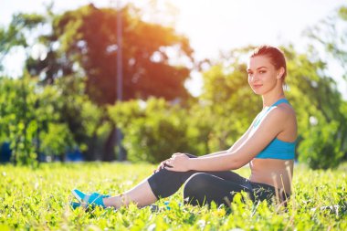 Aktif kıyafetli güzel bir kız güneş doğarken parkta dinleniyor. Yeşil çimlerin üzerinde oturan çekici genç bir kadının portresi. Açık havada eğitim ve meditasyon. Sabah egzersizleri ve sağlıklı yaşam tarzı..