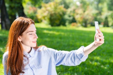 Genç kızıl saçlı kadın yeşil alanda selfie çekiyor. Akıllı telefonlu çekici bir kızın portresini yakından çek. Güneşli yaz gününde bahar parkında eğlenen bir kadın. Dijital mobil teknoloji