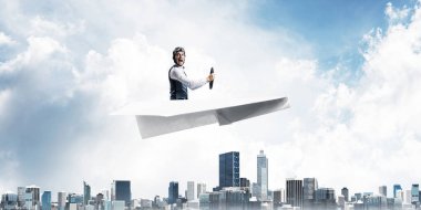 Kağıt uçakta oturan pilot ile iş liderliği ve motivasyon konsepti. Havacı şapkalı komik adam ve şehrin üzerinde kağıt uçak süren gözlükler. Yüksek binalarile modern megapolis.