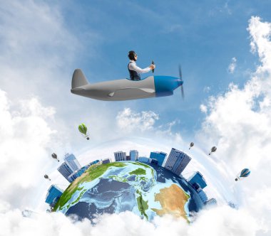 Pervane uçağı kullanan gözlüklü havacı şapkalı genç bir adam. Uçak konsepti ile dünyayı dolaşmak. Bulutlarla gökyüzünde küçük bir uçakla uçan komik adam. Gökdelenler ile yuvarlak toprak.