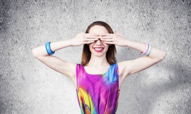 Kızıl saçlı güzel kız elleriyle gözlerini kapatıyor. Çekici bayan parlak elbiseler giyer ve gri duvar arkasında bilezikler takar. Hoş bir sürpriz bekleyen mutlu genç bir kadının portresi..