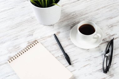 Bir fincan kahveyle ofis çalışma alanı. Düz tahta masa, açık spiral defter, gözlük ve kalemle. Kahve molası. Notlar için boş defter sayfası. Çaydanlıkta yeşil bitki.
