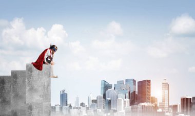 Maskeli ve pelerinli küçük üzgün çocuk binanın kenarında oturuyor ve aşağıya bakıyor. Karışık ortam