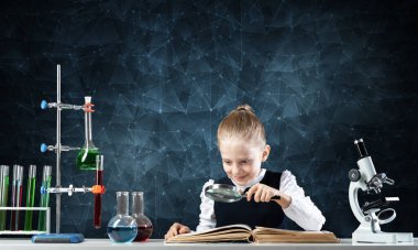 Küçük kız elinde büyüteçle masada oturuyor. Okulda araştırma ve eğitim. Okuldaki mutlu liseli kız kitap çalışırken dikkatli giyiniyor. Büyüteçle kitap okuyan zeki kız..