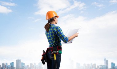 Teknik planla birlikte sabit şapkalı kadın mühendis. Kareli mavi gömlekli kadın mimarın şehrin arka planındaki inşaatı teftiş edişinin yan görüntüsü. Proje yönetimi inşa ediliyor