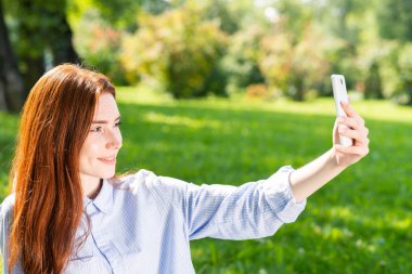 Genç kızıl saçlı kadın yeşil alanda selfie çekiyor. Akıllı telefonlu çekici bir kızın portresini yakından çek. Güneşli yaz gününde bahar parkında eğlenen bir kadın. Dijital mobil teknoloji