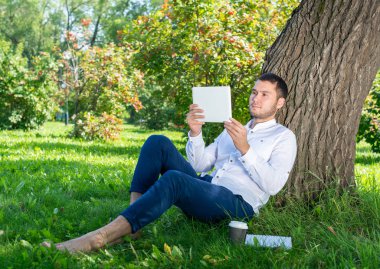 Güneşli bir günde parkta ağacın altında tablet bilgisayar kullanan genç bir adam. Tablet ekranına bakan yakışıklı bir iş adamı. Ağaca yaslanmış yeşil çimlerde oturan sakin bir adam. Mobil iletişim konsepti.