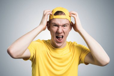 Stresli bir genç öfkeyle bağırıyor ve ellerini başının üstünde tutuyor. Duygusal kızıl oğlanın yüzünde tahriş olmuş bir ifade var. Adamın portresi gri arka planda sarı tişört ve beyzbol şapkası giyiyor.