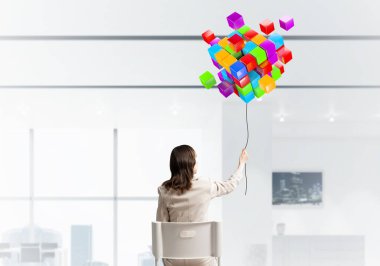 İple tutulan kadın 3D küplerden soyut renkli kompozisyon. Beyaz takım elbiseli uzun saçlı kadın, açık ofiste beyaz sandalyede oturuyor. Dijital teknoloji ve yenilik çözümleri.