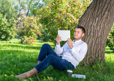 Güneşli bir günde parktaki ağacın altında tablet bilgisayar kullanan bir adam. Tablet ekranına bakan yakışıklı adam. Ağaçlara yaslanmış yeşil çimlerde oturan mutlu adam. Dijital teknoloji ve iletişim