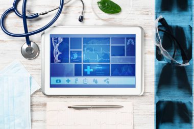 Doktorların ofisinde laboratuvar hasta muayenesi. Ekranda tıbbi uygulama arayüzü olan tablet bilgisayar. Stetoskop, röntgen görüntüsü ve kardiyogram tahta masada. Tıbbi teşhis ve muayene