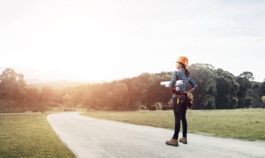 Kasklı genç bir kadın asfalt yolda elinde planlarla dikiliyor. İş kıyafetlerindeki kadın mimarın kırsal kesimdeki yeni inşaat alanlarını kontrol edişinin arka görüntüsü. Yol altyapısı yeniden yapılandırması.
