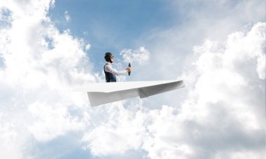 Havacı şapkalı işadamı kağıt düzlemde oturuyor ve direksiyonu tutuyor. Bulutlu mavi gökyüzünde kağıt uçak kullanan pilot. Ekstrem havacılık hobisi. Kabarık bulutlu gökyüzü panoraması.