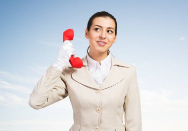 Kırmızı retro telefonu tutan çekici genç bir kadın. Beyaz takım elbiseli çağrı merkezi operatörü gökyüzü arka planında telefonla poz veriyor. Tele-pazarlama hattı. İş yardımı ve danışma.