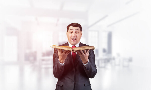 Surprised businessman holding book with open mouth. Amazed adult man in business suit and tie standing in blurred office interior. Education and knowledges. Professional business accounting service