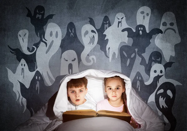 害怕的女孩和男孩在床上读孩子的书 惊慌失措的孩子一起躺在毯子下 穿着睡衣和滑稽鬼的孩子在灰墙上剪影 儿童阅读魔法童话 — 图库照片
