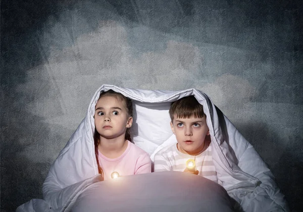 被吓得的孩子 用手电筒躺在床上 小妹妹和弟弟一起躲在毯子下 被覆盖的孩子害怕晚上在灰色墙壁的背景黑暗 儿童夜惊 — 图库照片