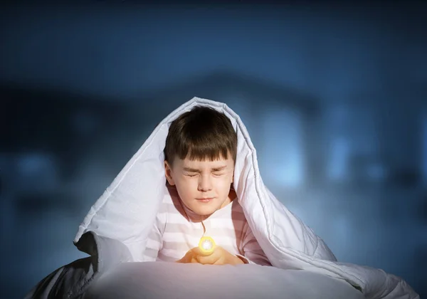 懐中電灯を毛布の下に隠れて恐怖の子供 家のベッドに寝そべって目を閉じて怖がっている子供 暗い概念の恐怖 深い青の背景にパジャマの男の子の肖像画 — ストック写真