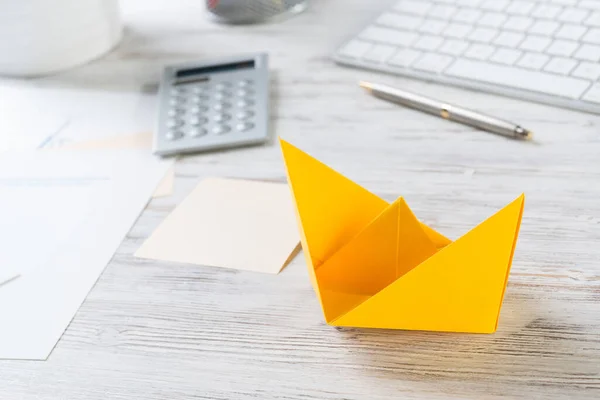 Businessman workspace with office wooden desk and yellow paper ship. Flat lay table with calculator and business documents. Close up orange origami boat. Creative and innovative solution for business.
