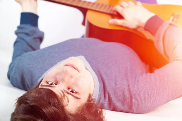 Young man lying back on sofa with guitar. Handsome guitarist relaxing on white sofa at home. Portrait of musician playing acoustic guitar. Freetime activity and playing musical instrument